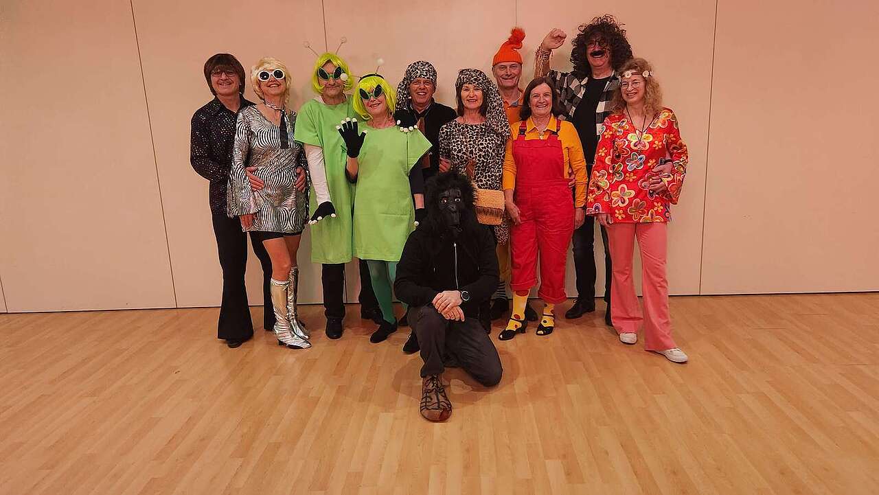 Eine Gruppe von Tänzern posiert in verschiedenen Mottokostümen, darunter Hippie, Disco und Alien
