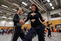 Hilde und Thomas Schütze tanzen energiegeladen Discofox in der Sporthalle; Hilde im schwarzen Glitzer-Outfit, Zuschauer im Hintergrund.